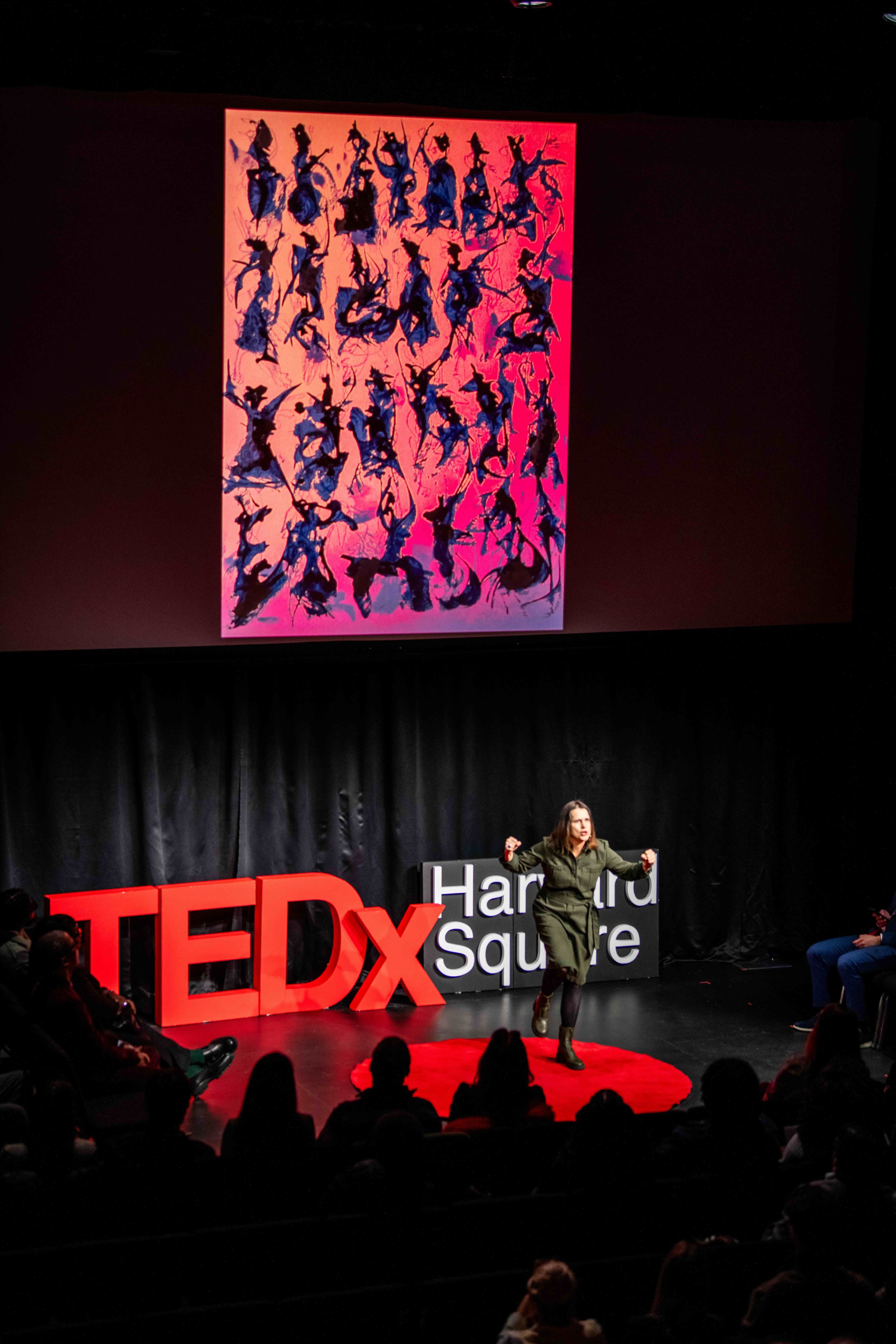 A speaker performs on the TEDxHarvardSquare stage beneath a large projected artwork of dancing silhouettes.