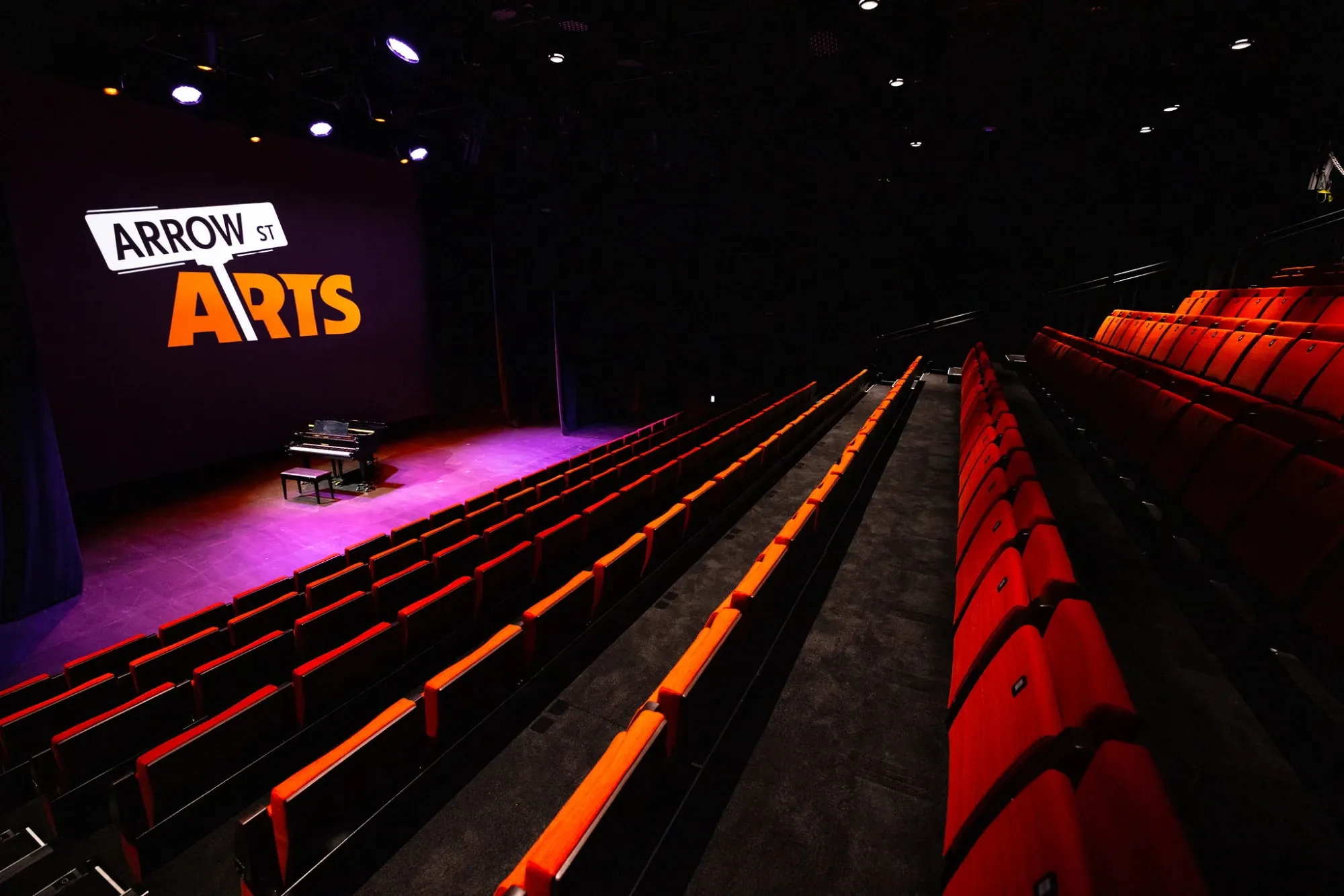 The empty Arrow Street Arts theater: rows of red seats rise toward a stage with a grand piano, under the venue's projected sign.