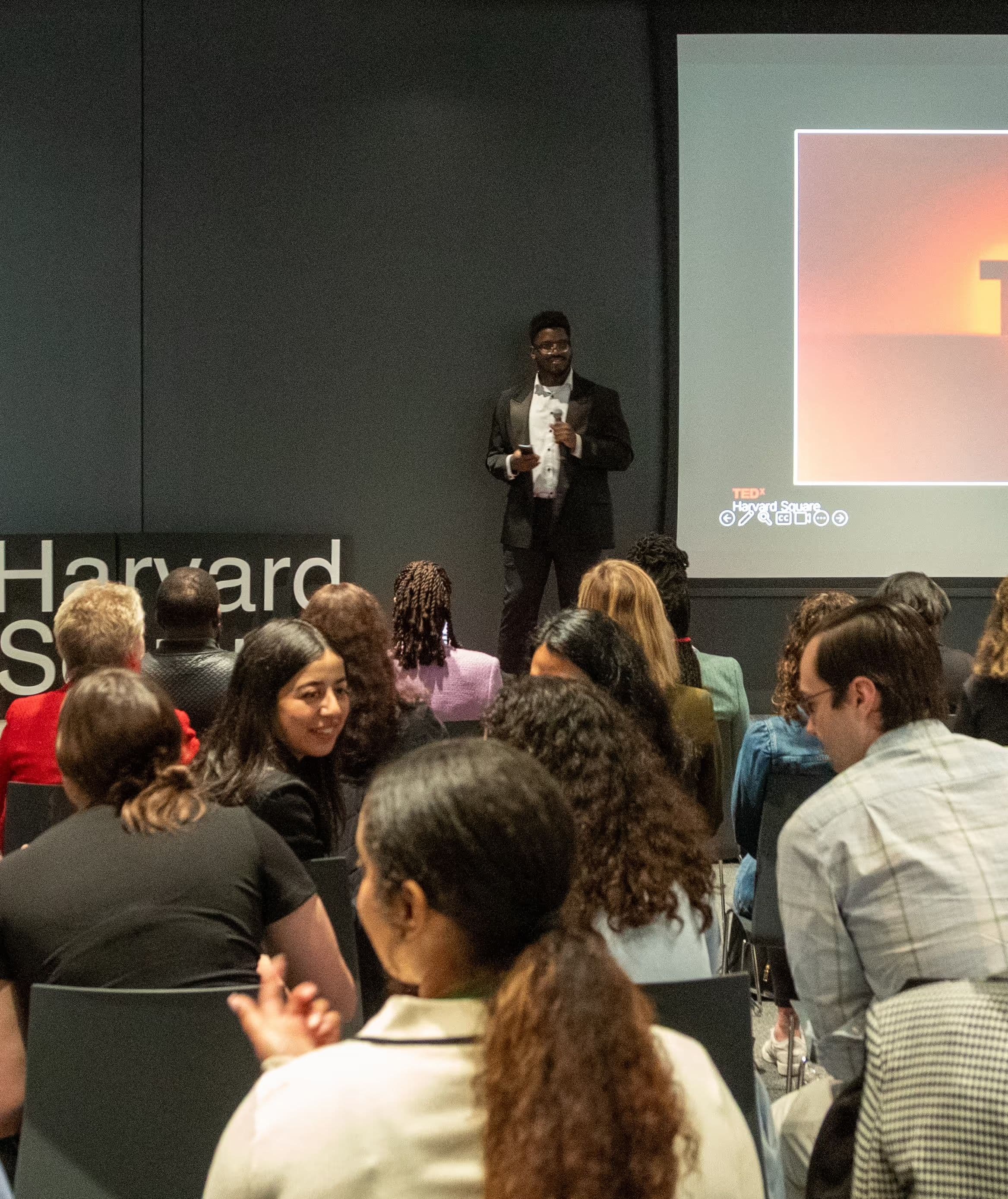A speaker in a black suit addresses a seated audience in a gallery-style room, with the Harvard Square wordmark and a projected slide behind him.